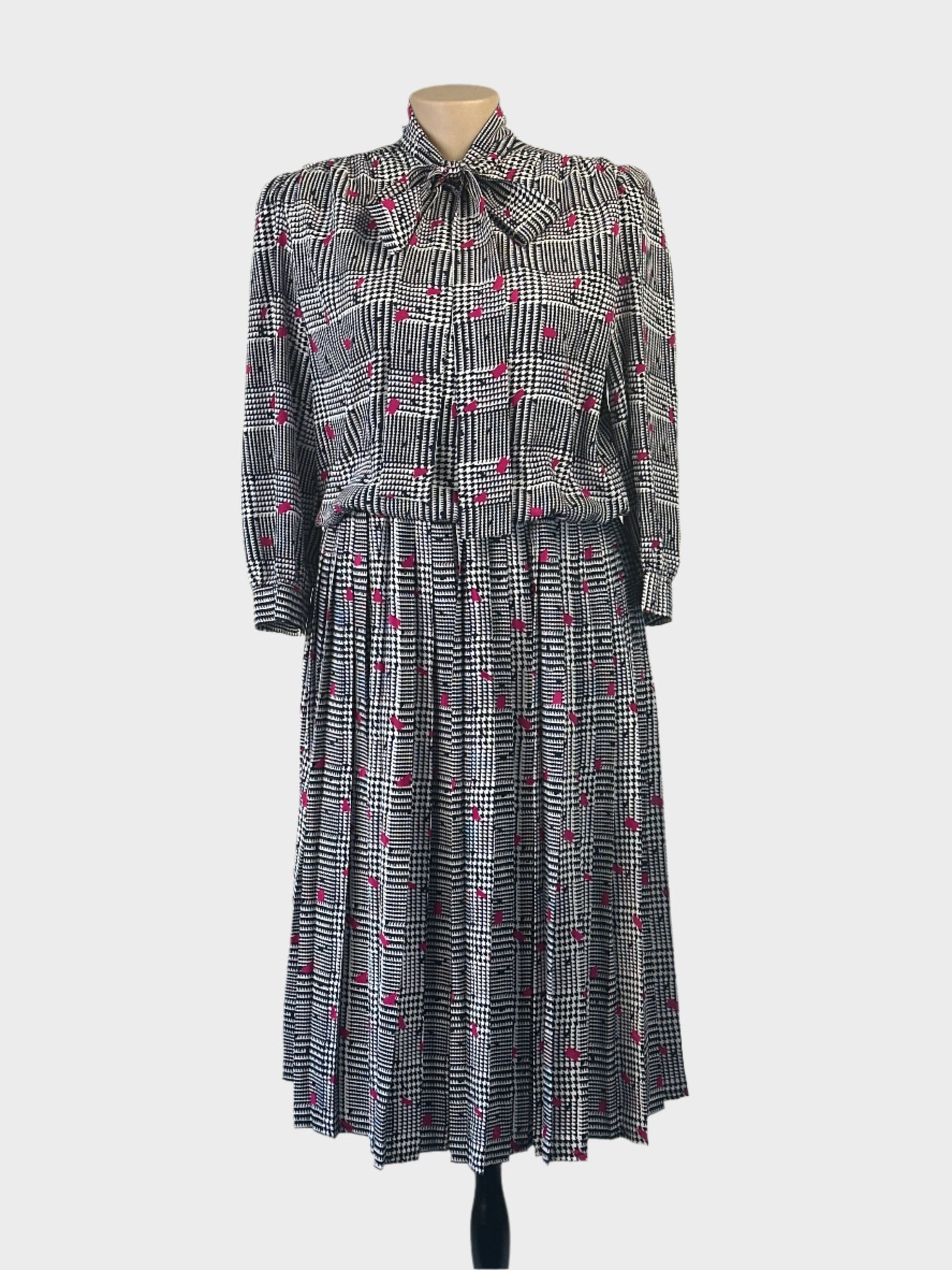 Vintage 80’s Noel Wells blouson dress in houndstooth check with contrasting flecks, band collar with pussy bow tie, and pleated skirt for office or special events.